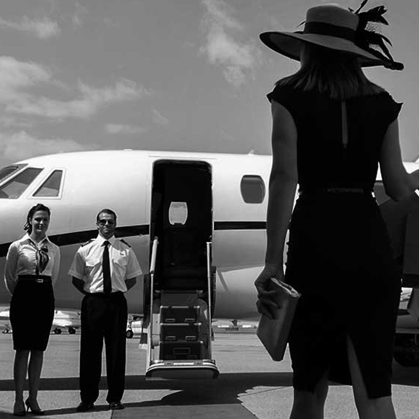 Black-and-white photograph of a traveller approaching a private jet, with two uniformed crew members beside the boarding stairs.