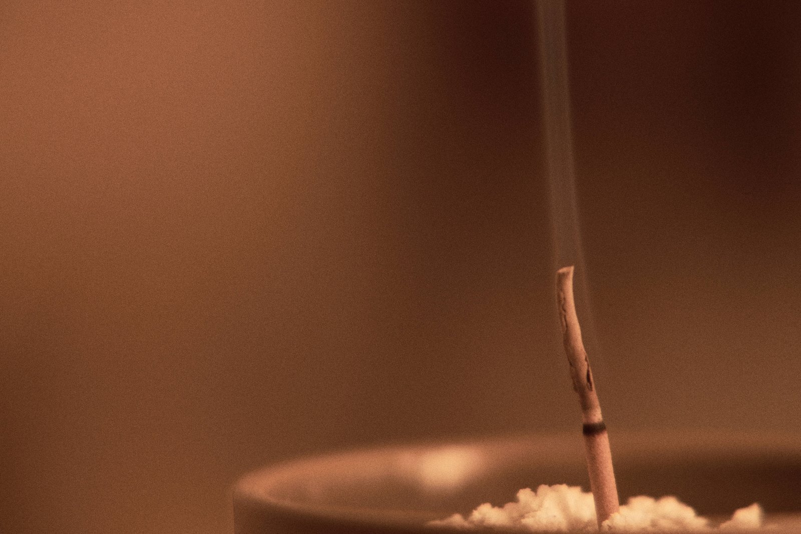 An incense stick releasing a fine trail of smoke above a dark incense dish against a softly blurred warm brown background.