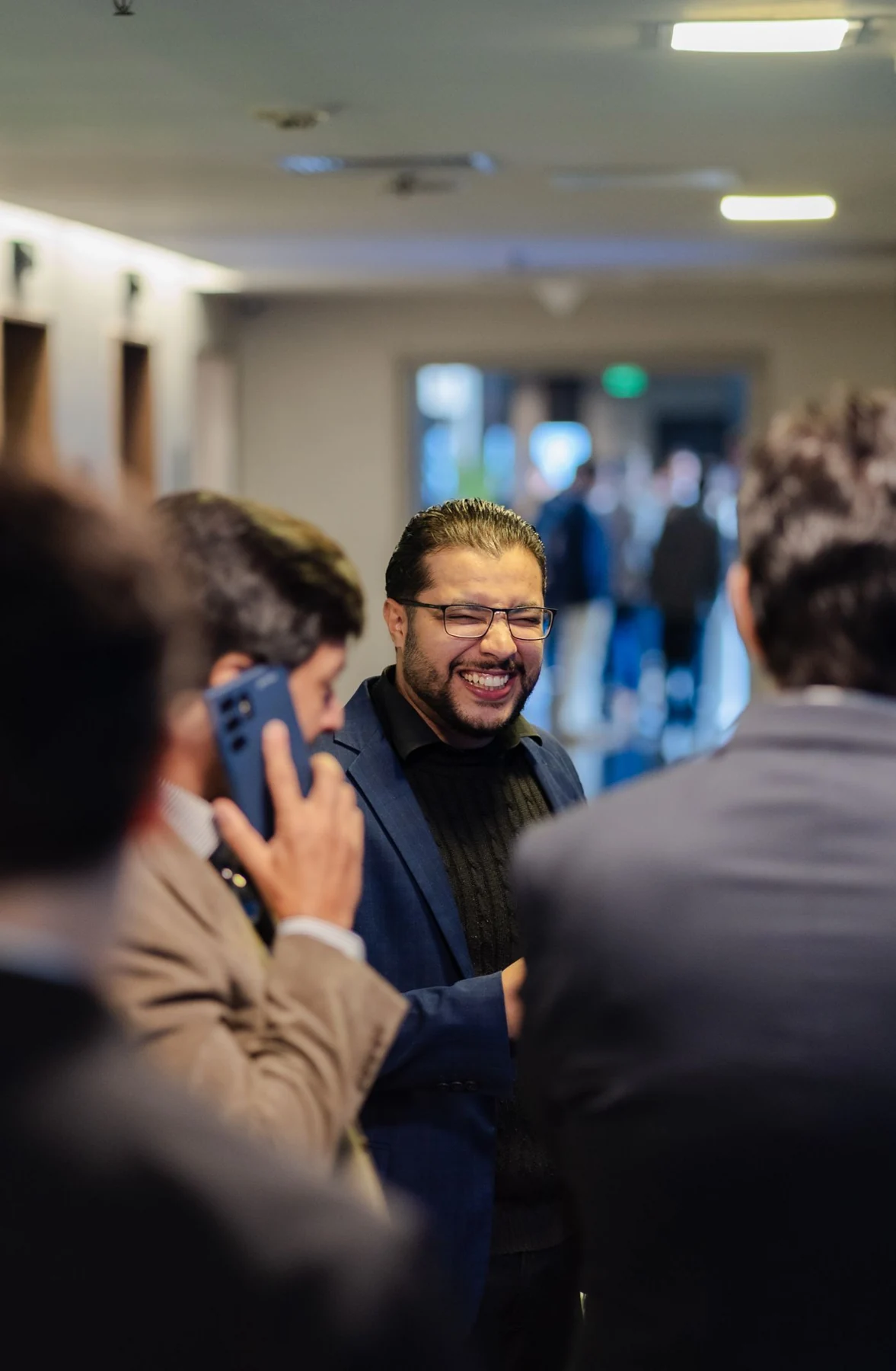 Business professionals converse at an indoor networking event, with a smiling attendee in focus and others softly blurred in the foreground.