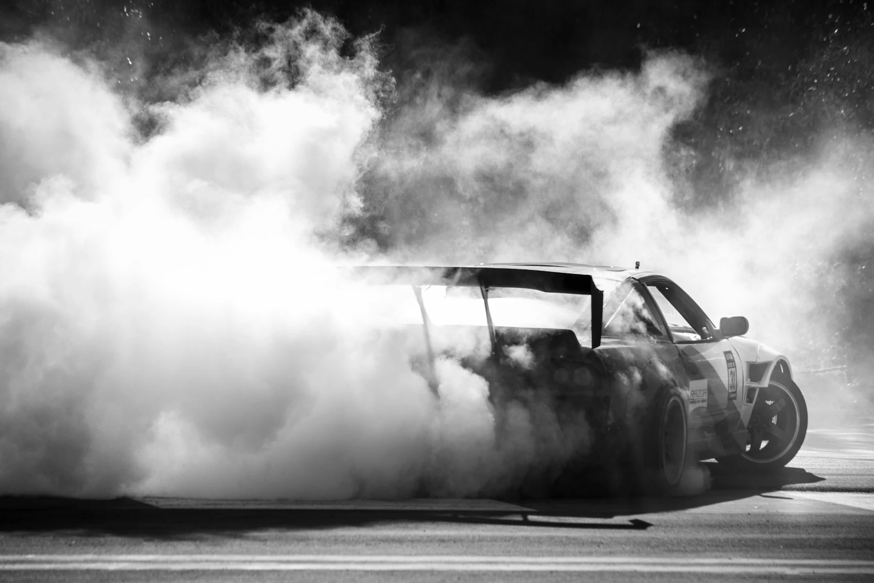 A monochrome photograph of a drift car with a large rear wing surrounded by tyre smoke on a track.