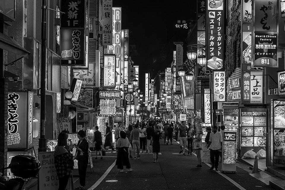 Black-and-white photograph of pedestrians beneath illuminated neon signs on a Kabukichō street in Shinjuku, Tokyo, at night.