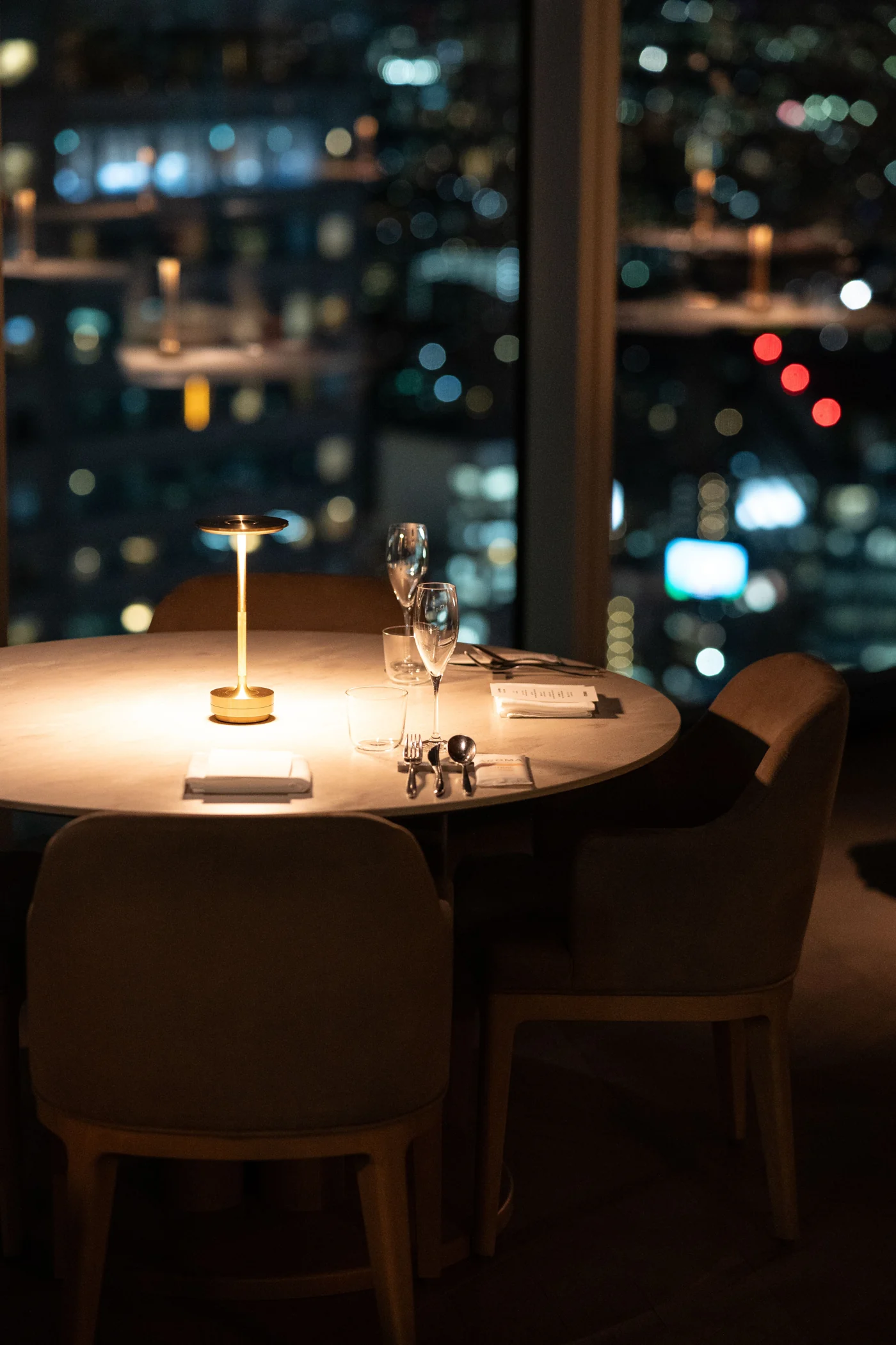 An elegant round dining table with a small gold lamp, glassware and brown chairs beside a window overlooking Tokyo at night.